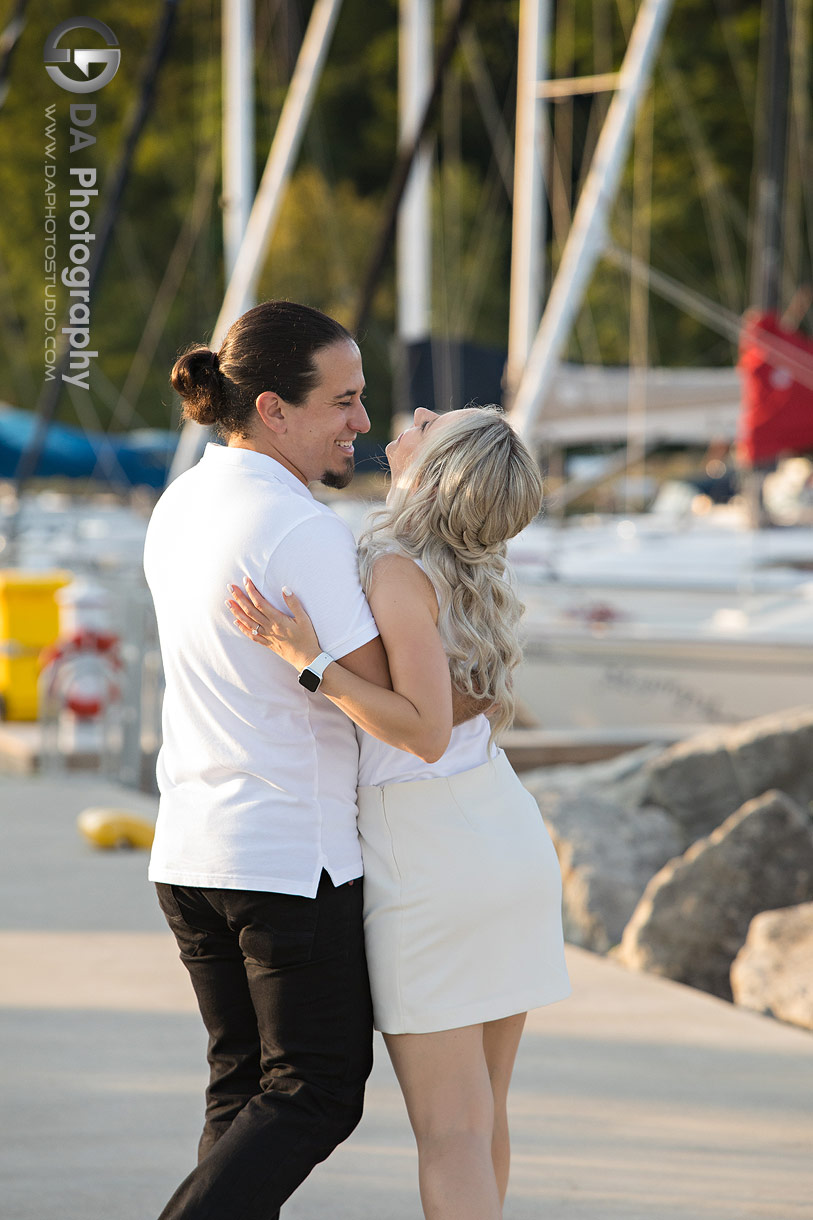 Engaged couple embracing in a shaded area overlooking boats at the marina